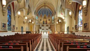 Interior of an ornate church with vaulted blue and beige ceilings, chandeliers, and rows of wooden pews leading to a white altar.