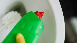 Close-up of a green squeeze bottle with a red nozzle dispensing liquid toward a white plate in the background.