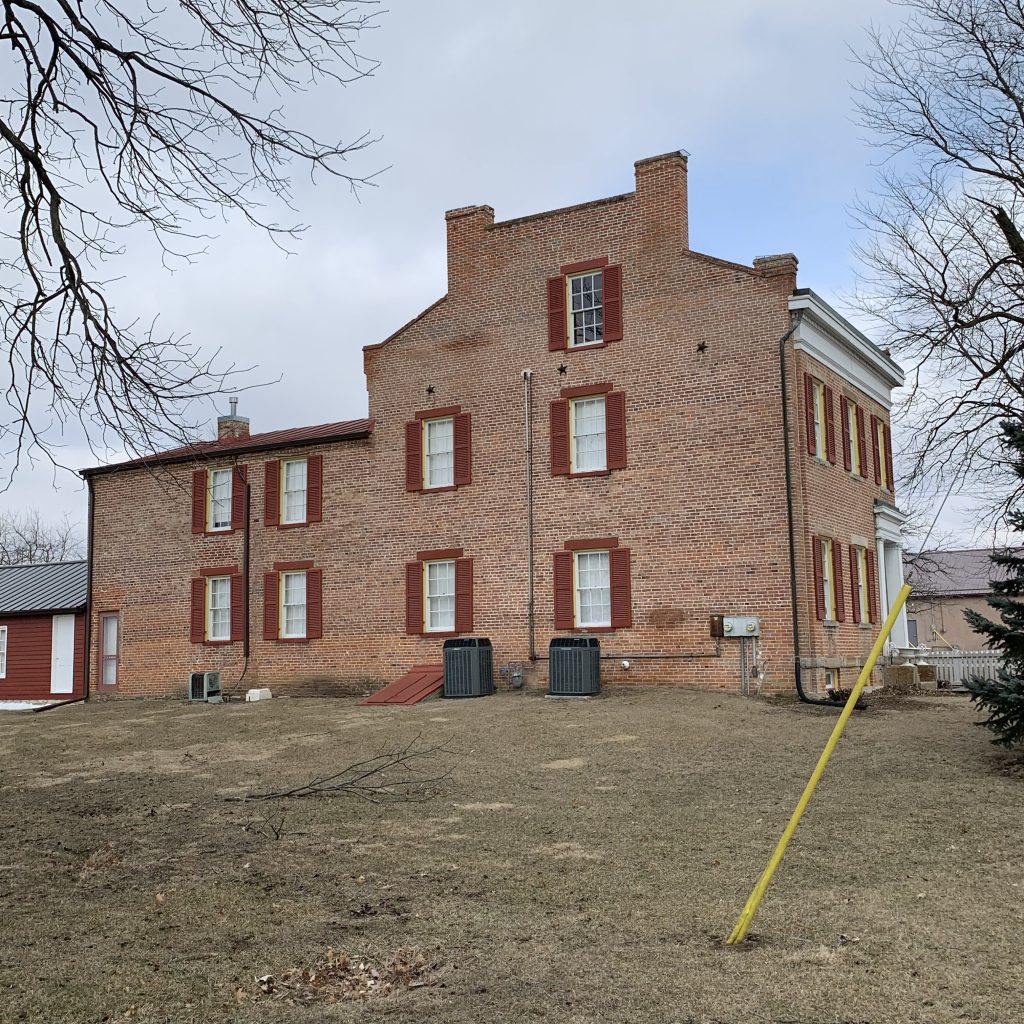 Brick building with red shutters on a wintery, leafless-tree street and an empty dirt lot in front.