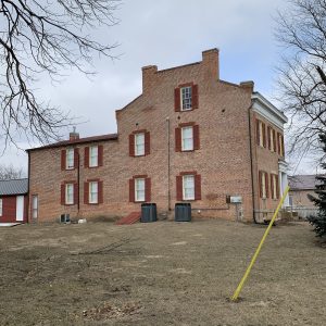 Brick building with red shutters on a wintery, leafless-tree street and an empty dirt lot in front.