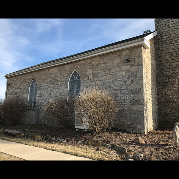 Stone church side with two tall arched windows, beige stone walls, and bare landscaping under a blue sky.
