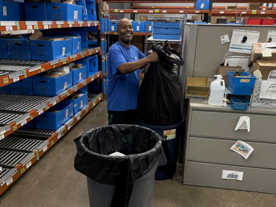 Man in a blue shirt standing in a storage aisle, placing a black trash bag into a large trash can among blue storage bins and office supplies.